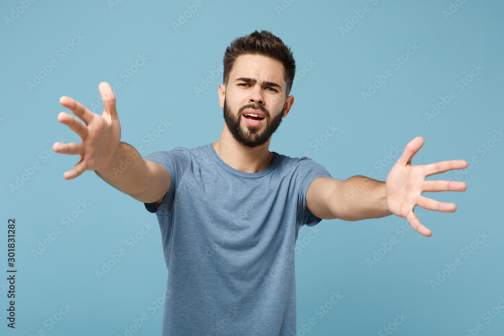 Fototapeta premium Young perplexed displeased man in casual clothes posing isolated on blue background, studio portrait. People sincere emotions lifestyle concept. Mock up copy space. Standing with outstretched hands.