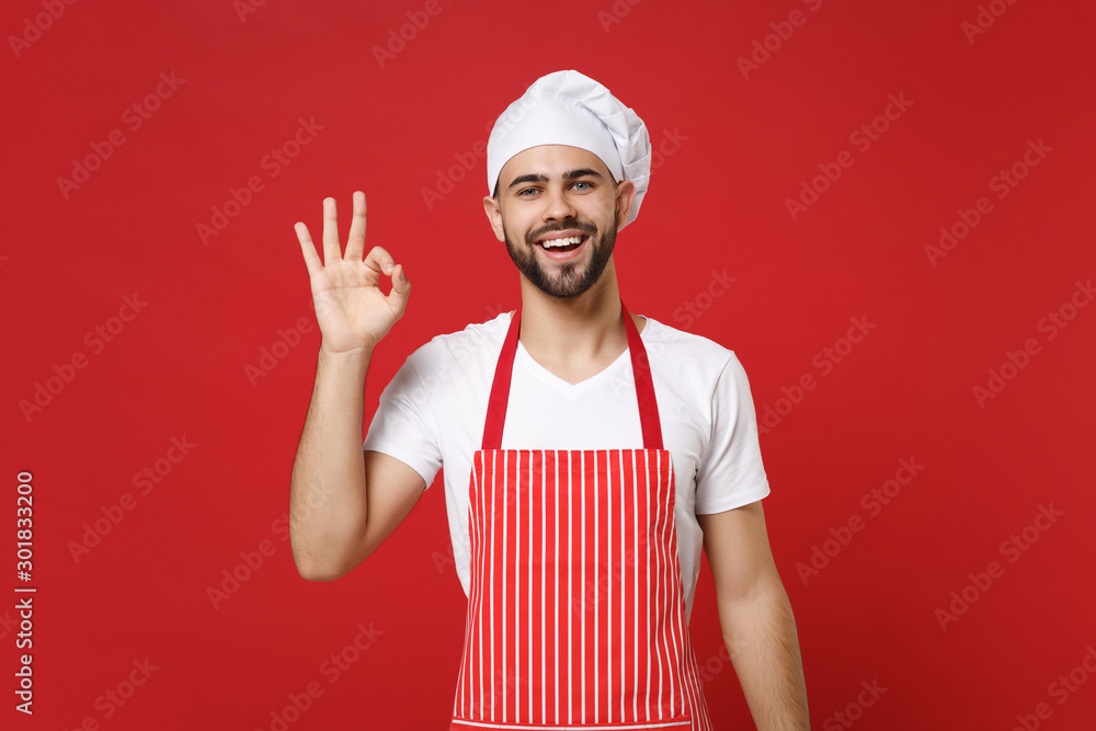 Smiling young bearded male chef cook or baker man in striped apron white t-shirt toque chefs hat posing isolated on red wall background. Cooking food concept. Mock up copy space. Showing OK gesture. Smiling young bearded male chef cook or baker man in striped apron white t-shirt toque chefs hat posing isolated on red wall background. Cooking food concept. Mock up copy space. Showing OK gesture.