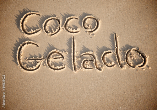 'Coco Gelado' (Translation: Chilled coconut water) handwritten text in the sand of a beach in Brazil