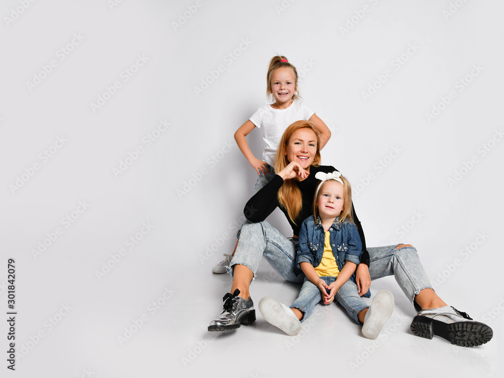 Happy young mother is sitting on the floor posing with two daughters preschoolers on white. Human relations, family. Happy young mother is sitting on the floor posing with two daughters preschoolers on white. Human relations, family.