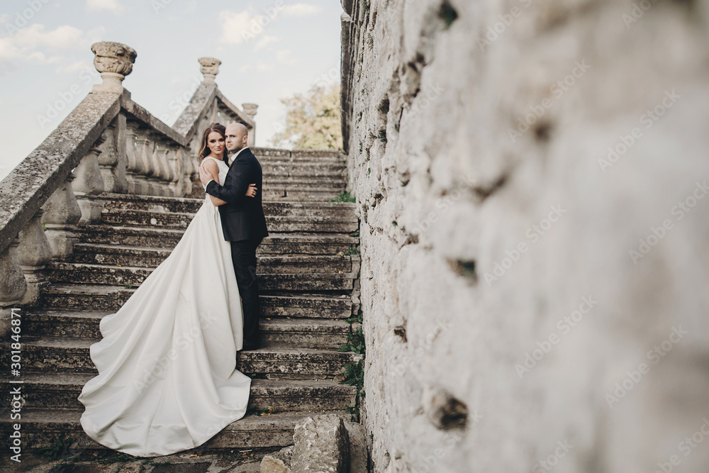 Gorgeous wedding couple walking on stone stairs near old castle in park. Stylish beautiful bride in amazing gown and  groom posing on background of ancient building