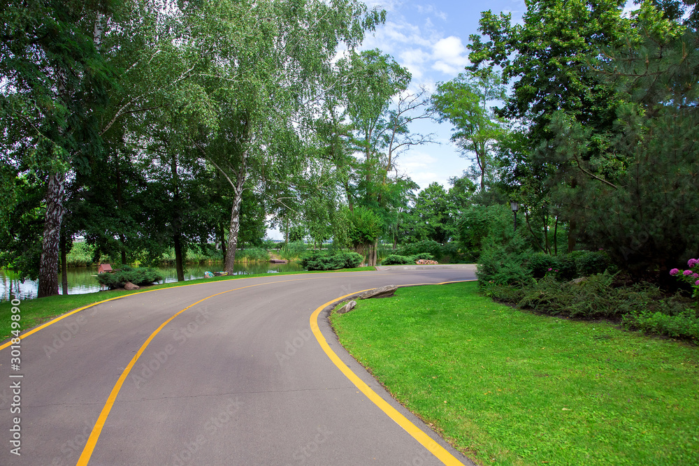 asphalt road with yellow markings leaving for a turn in the park with a green lawn and trees on the side of the road and a river with a blue sky in the background. asphalt road with yellow markings leaving for a turn in the park with a green lawn and trees on the side of the road and a river with a blue sky in the background.