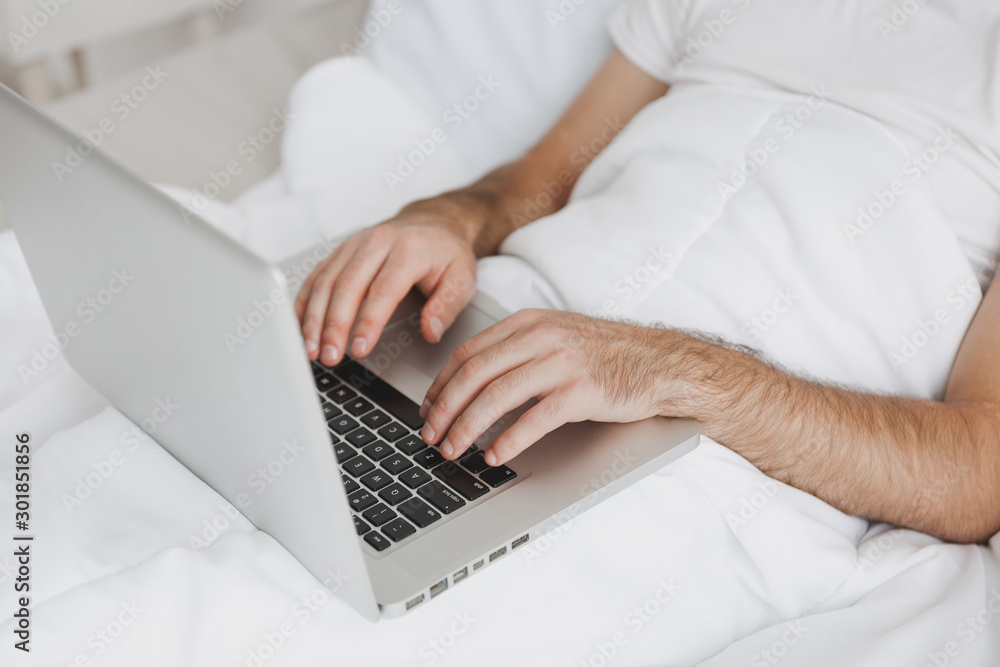 Fototapeta premium Close up cropped image of man lying in bed with white blanket in bedroom at home. Male spend time in room using pc laptop. Hands on keyboard. Rest relax good mood lifestyle concept. Mock up copy space