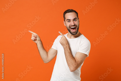 Crazy young man in casual white t-shirt posing isolated on bright orange wall background studio portrait. People sincere emotions lifestyle concept. Mock up copy space. Pointing index fingers aside.