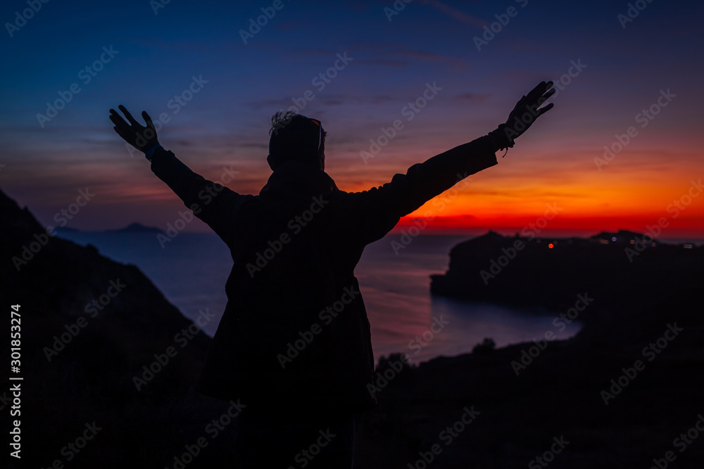 Man traveler silhouette hiking at sunset on Santorini island. Happy tourist raised arms feeling free, admiring landscape