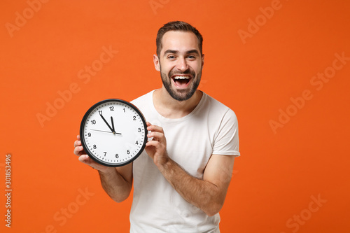 Cheerful young man in casual white t-shirt posing isolated on bright orange wall background studio portrait. People sincere emotions lifestyle concept. Mock up copy space. Holding in hand round clock.