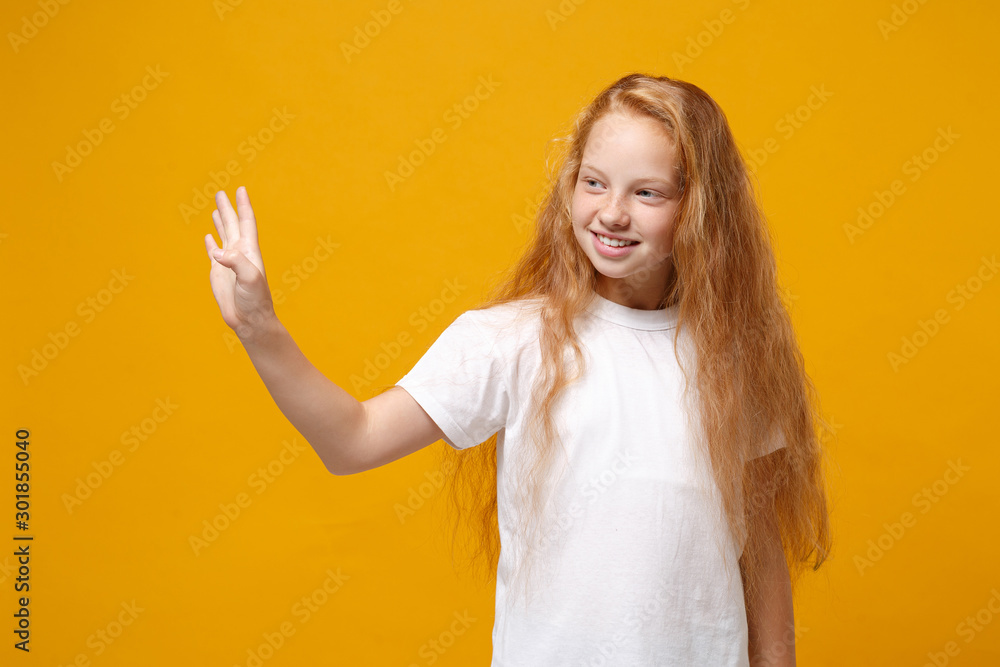 Smiling little ginger kid girl 12-13 years old in white t-shirt isolated on bright yellow background. Childhood lifestyle concept. Mock up copy space. Waving and greeting with hand as notices someone. Smiling little ginger kid girl 12-13 years old in white t-shirt isolated on bright yellow background. Childhood lifestyle concept. Mock up copy space. Waving and greeting with hand as notices someone.