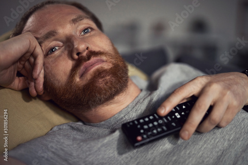 Fotografie Portrait of bearded adult man watching TV at night while lying on couh in dark r