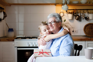 Child is giving gift box to grandmother. Kid and senior woman are hugging in cozy home kitchen. Happy family is enjoying kindness. Lifestyle moment. Concept of holidays Hanukkah, Christmas, Birthday.