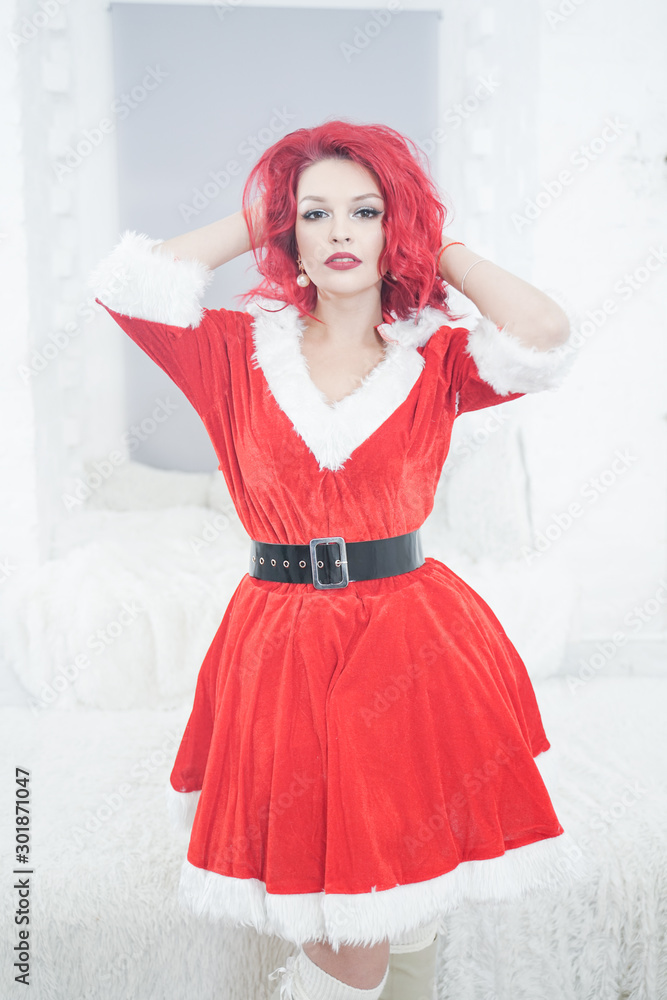 pretty redhaired young stylish woman in velvet santa dress with fur posing on white room background near the cozy window pretty redhaired young stylish woman in velvet santa dress with fur posing on white room background near the cozy window