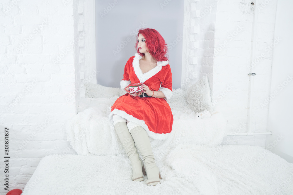 Fototapeta premium pretty cheerful happy redhaired adult girl ready for christmas and enjoy by rest with big cup near her window sill with fur pillows alone in white room