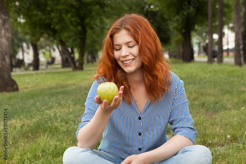 Young red-haired girl eating a green apple in the park.