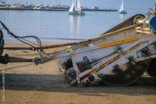 A special machine filters sand and removes waste and seaweed from it on blue sea and sailboats background. Beach garbage cleaning machine at work. Catalonia, Spain. Preparing the beach for the summer.