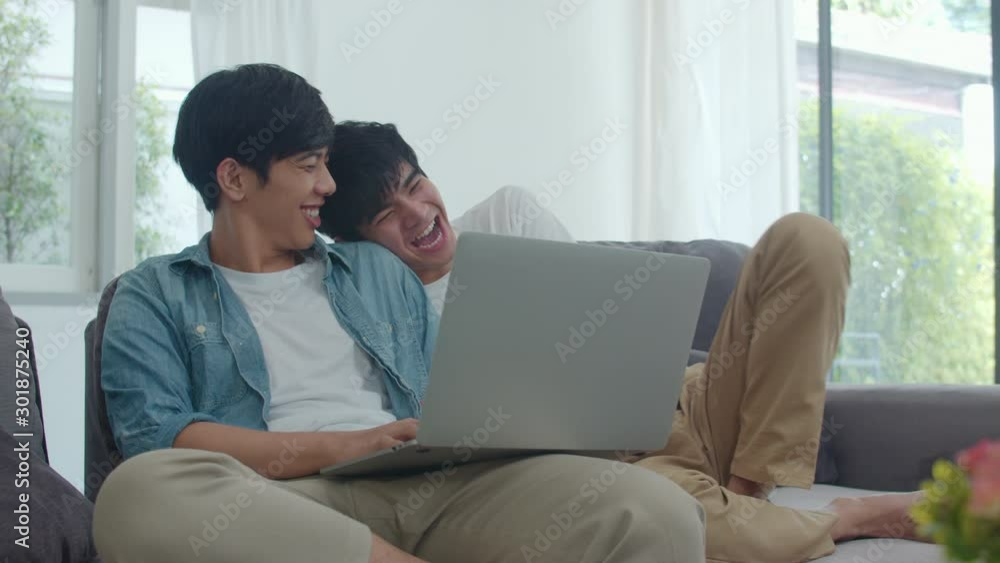 Young Gay couple using computer laptop at modern home. Asian LGBTQ men happy relax fun using technology watching movie in internet together while lying sofa in living room at house concept. Young Gay couple using computer laptop at modern home. Asian LGBTQ men happy relax fun using technology watching movie in internet together while lying sofa in living room at house concept.