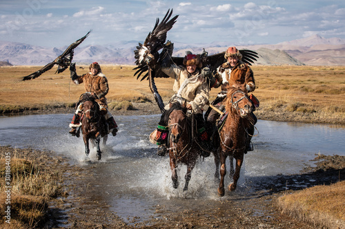 Fotografiet A group of traditional kazakh eagle hunters holding their golden eagles on horseback while galloping through a river