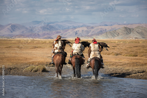 A group of traditional kazakh eagle hunters holding their golden eagles on horseback while walking through a river. Ulgii, Mongolia.