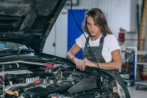 Photo of young girl car repair worker.Beautiful brunette wearing black jumpsuit. Girl staying near the car in car servise working under car cowl.