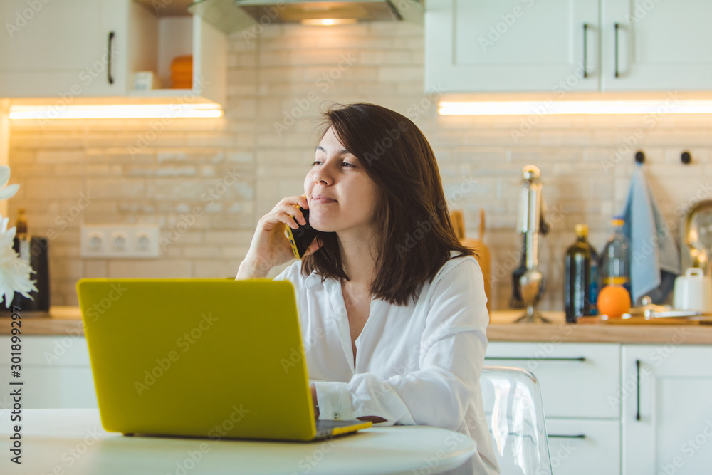 young pretty caucasian woman talking on the phone sitting in front of laptop in the kitchen young pretty caucasian woman talking on the phone sitting in front of laptop in the kitchen
