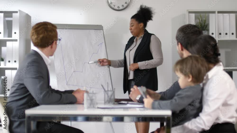African american businesswoman showing graph on flipchart and giving presentation to colleagues during office meeting while little boy sitting on knees of mother and playing on digital tablet African american businesswoman showing graph on flipchart and giving presentation to colleagues during office meeting while little boy sitting on knees of mother and playing on digital tablet