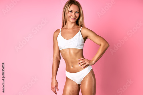 Active sportive woman with long natural hair, wearing nice white swimsuit, looking straight at camera with engaging smile, expressing positiveness and joyfulness, keeping hand on tight hips