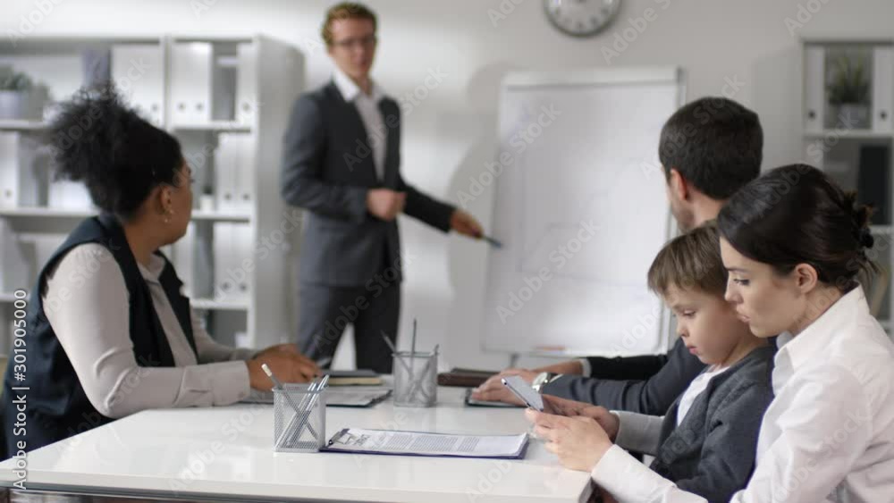 Businesswoman sitting with little son on her knees and using digital tablet during office meeting with colleagues while businessman giving presentation on flipchart Businesswoman sitting with little son on her knees and using digital tablet during office meeting with colleagues while businessman giving presentation on flipchart