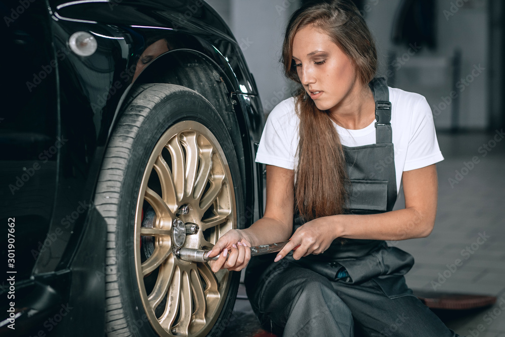 A beautiful brunette girl in a white T-shirt and black jumpsuit is sitting near a black car removing the wheel in the service station
