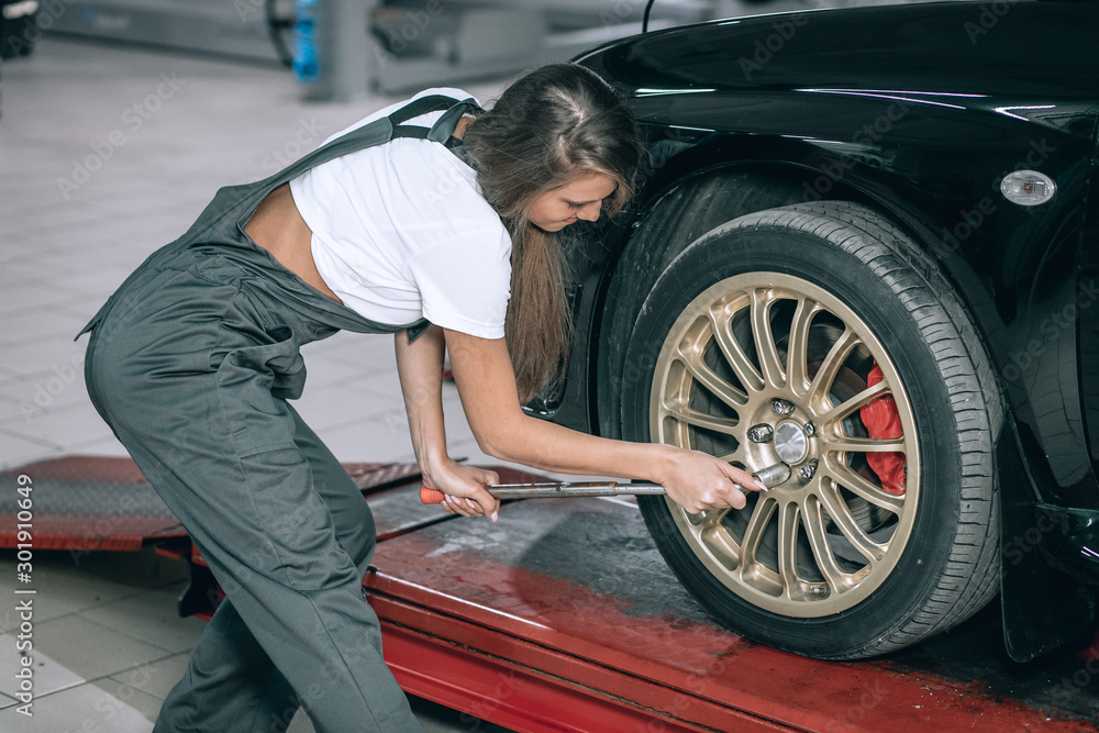 A beautiful girl in a white T-shirt and black jumpsuit is sitting near a black car removing the wheel in the service station A beautiful girl in a white T-shirt and black jumpsuit is sitting near a black car removing the wheel in the service station