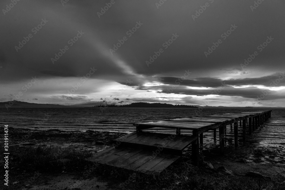 View of a pier on a lake, with a ray of sun against a moody sky