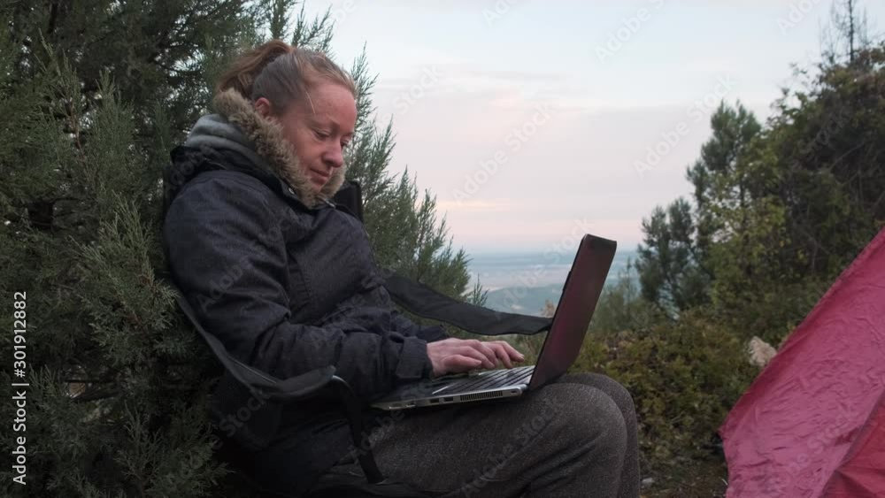 woman works on laptop while sitting on chair near tent on top of mountain. woman works on laptop while sitting on chair near tent on top of mountain.