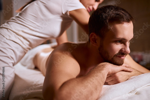 Close up of handsome man with thick stylish beard having aroma therapy massage with essential oil, looking away with satisfied expression, lying on white towels, shot from below