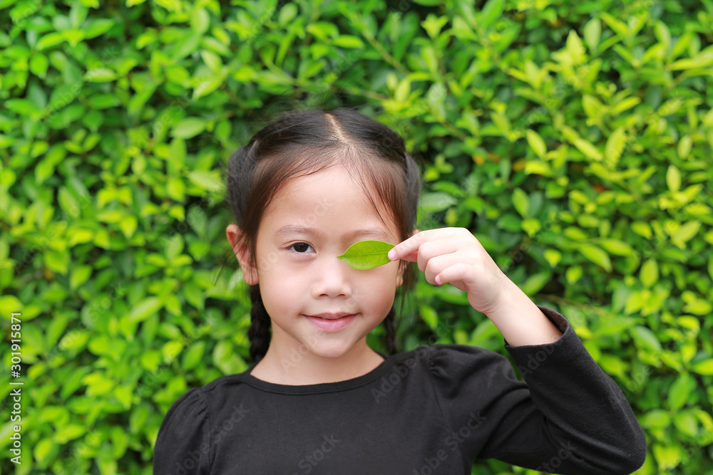 Cute little child girl holding a green leaf closing left eye in green garden background. Cute little child girl holding a green leaf closing left eye in green garden background.