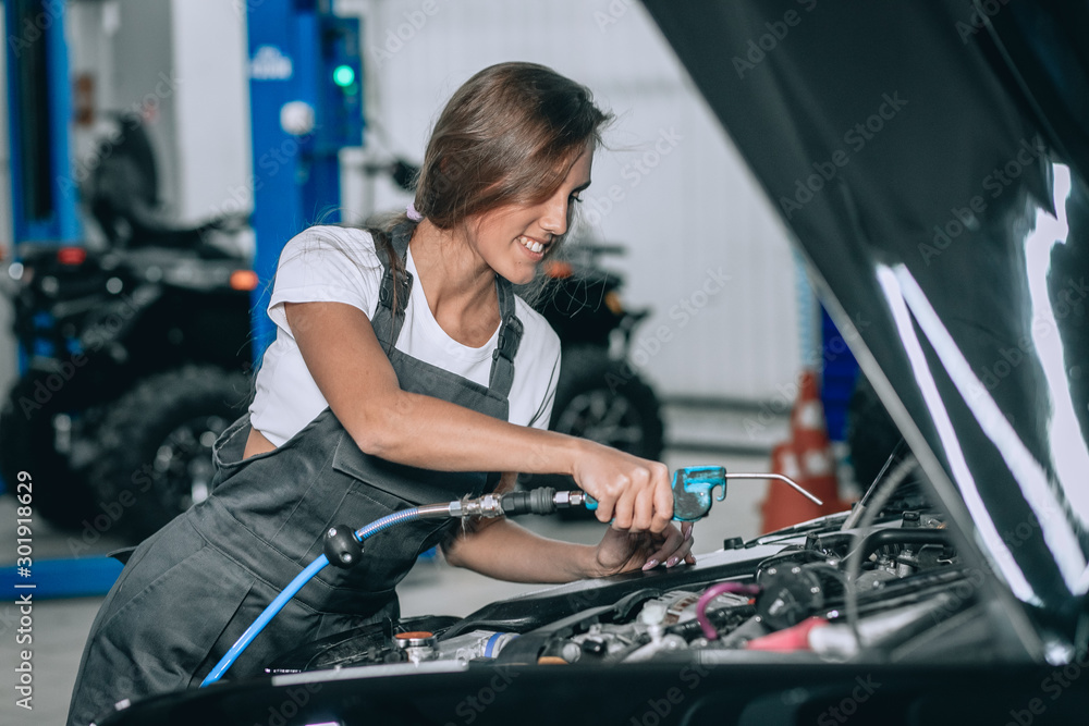 A beautiful girl in a black jumpsuit and a white t-shirt is smiling, checking the oil level in a black car in the garage.