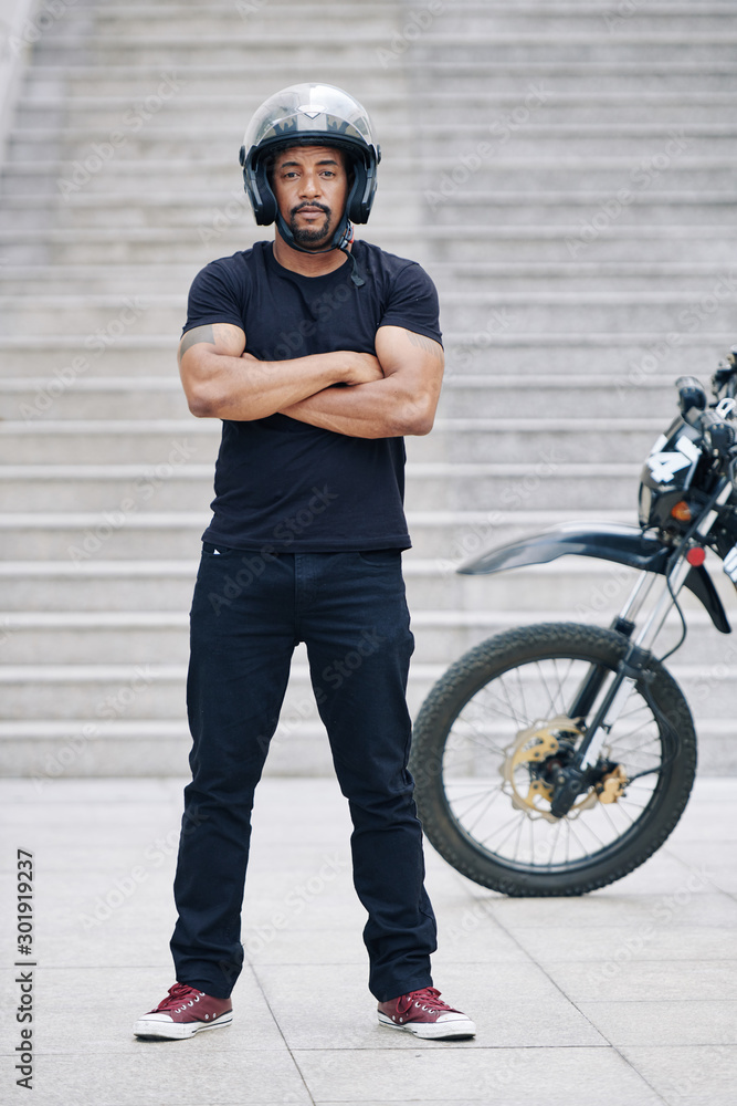 Handsome mixed-race muscular motorcyclist standing at stairs after riding through the city Handsome mixed-race muscular motorcyclist standing at stairs after riding through the city