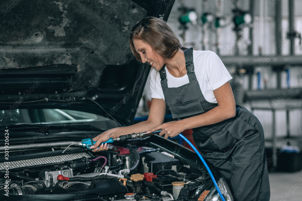 A beautiful girl in a black jumpsuit and a white t-shirt is smiling, checking the oil level in a black car in the garage.