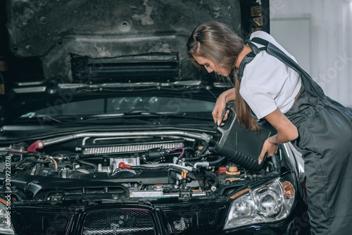 A beautiful girl in a black jumpsuit and a white t-shirt is smiling  checking the oil level in a black car in the garage.