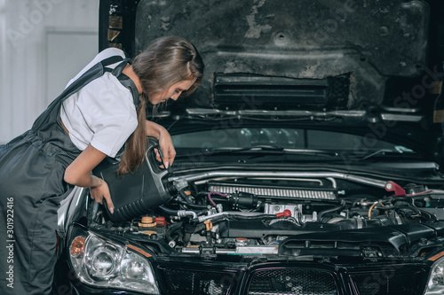 A beautiful girl in a black jumpsuit and a white t-shirt is smiling, checking the oil level in a black car in the garage.