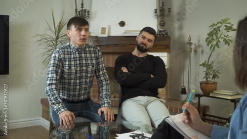Stressed Caucasian boy talking to the female psychologist white another bearded man sitting next to him and smiling sneeringly. Sarcastic guy came with friend at the psychological consultation. photo