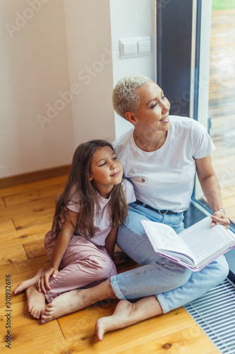 Beautiful mother and her daughter sit on floor reading book near big panoramic window. White colors