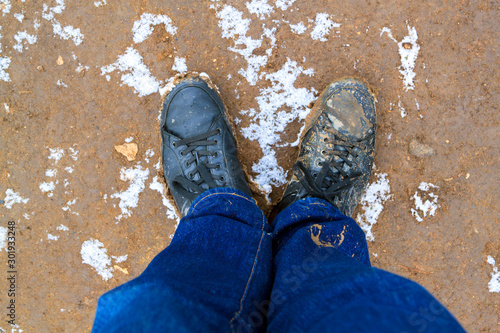 top view of black boots and legs in blue jeans all covered in mud standing on dirty ground covered with snow. Concept of leaving the comfort zone to meet new challenges
