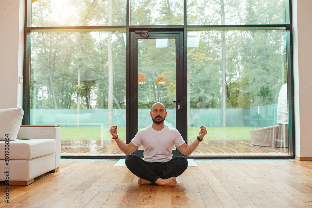 Male in casual clothes sit in lotus position meditate, relax. Background big panoramic window. Yoga at home concept