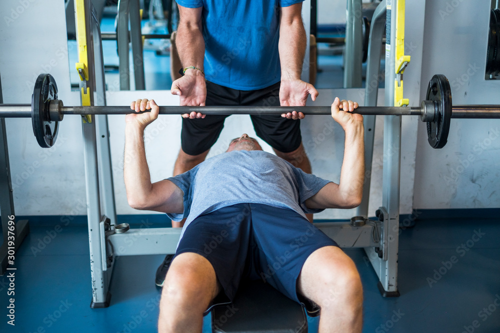 one mature man lying down on the bench of the gym training hus body to be fitness and active senior - man helping he holding de bar with his hands one mature man lying down on the bench of the gym training hus body to be fitness and active senior - man helping he holding de bar with his hands