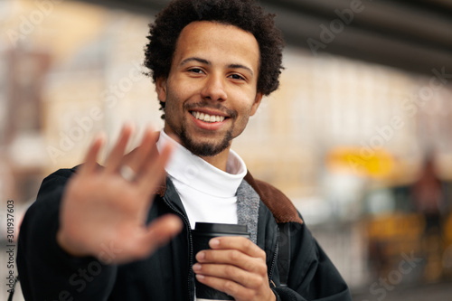 Portrait of African man with cup of coffee, raises his hand with gesture of greeting.