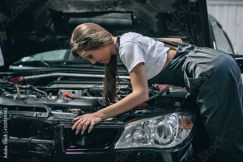 A brunette in a black jumpsuit and a white t-shirt near the open hood of black car. Young female in the garage is smiling at the camera and lowered gaze. car repair concept