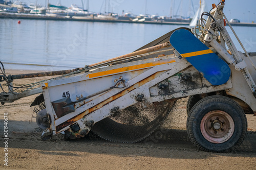 A special machine filters sand and removes waste and seaweed from it on blue sea background. Beach garbage cleaning machine at work. Roses, Catalonia, Spain.