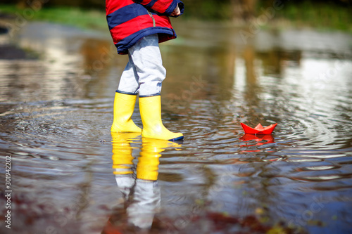 Happy little kid boy in yellow rain boots playing with paper ship boat by huge puddle on spring or autumn day. Active leisure for children. Funny child having fun outdoors, wearing colorful clothes.