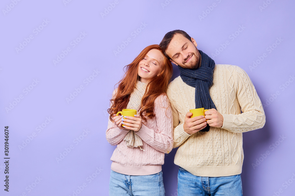 Romantic pleasant couple, drinking hot fresh tea, holding yellow cups, bowing heads towards each others, closing eyes with pleasure, sweet home concept, isolated on violet background