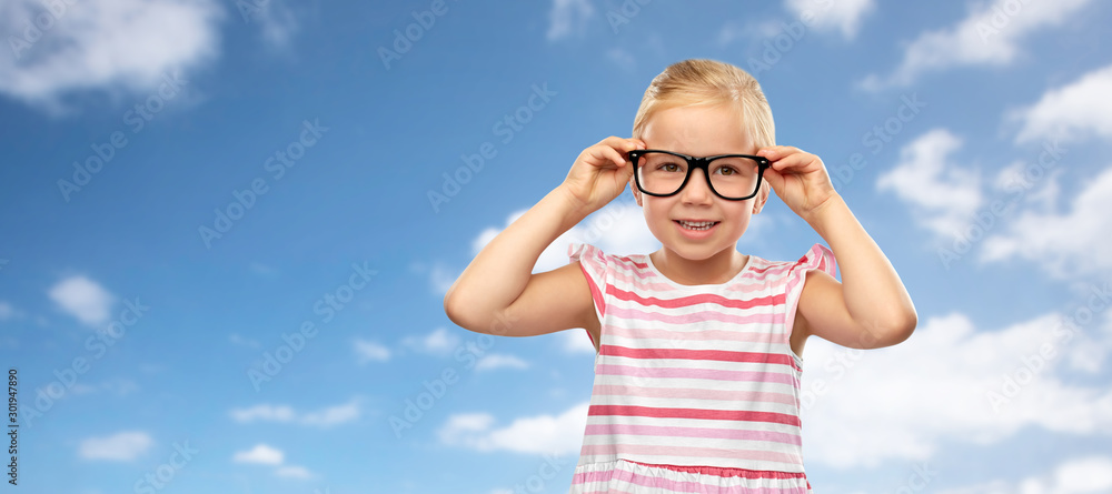 education, school and vision concept - smiling cute little girl in black glasses over blue sky and clouds background