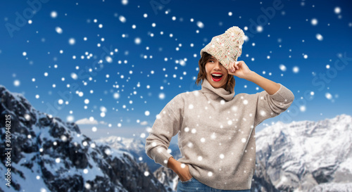 christmas, season and people concept - happy smiling young woman in knitted winter hat and sweater over snow and alps mountains background