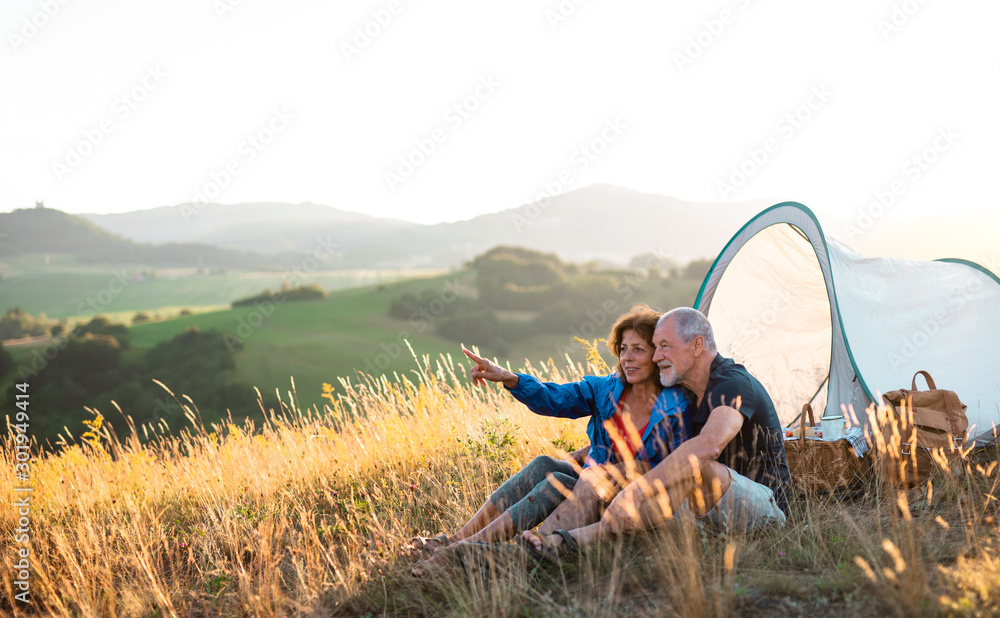 Senior tourist couple in love sitting in nature at sunset, resting. - obrazy, fototapety, plakaty 