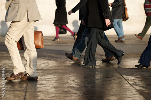 Unrecognizable commuters walking in bright sun across wet pavement in Trafalgar Square, London, UK